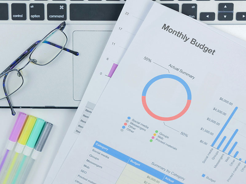 Monthly budget document with pie chart, bar graph, glasses on laptop, colorful markers.