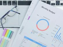 Monthly budget document with pie chart, bar graph, glasses on laptop, colorful markers.